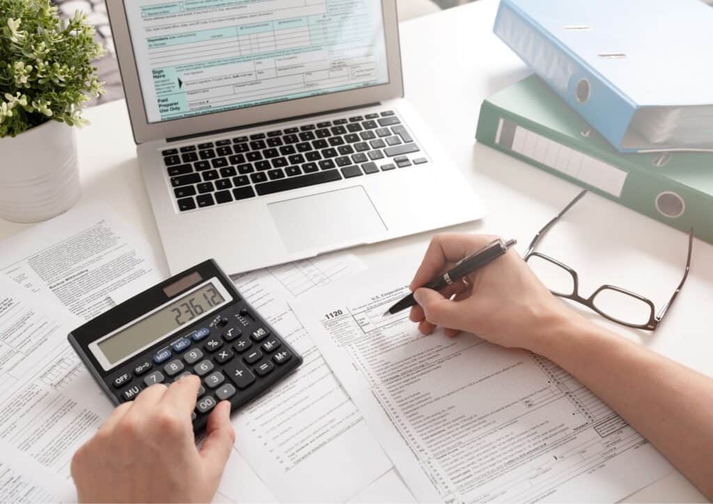 Person working with a laptop and filling in paperwork using a calculator.