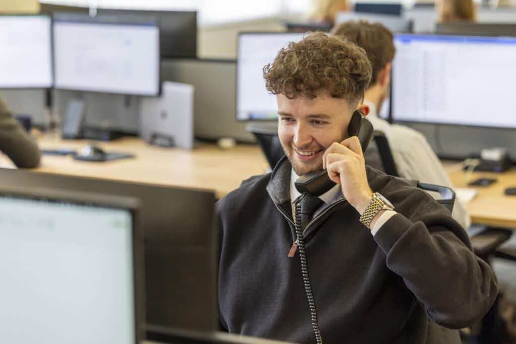 Matthew Crabtree from GLX Advisory wears a dark-coloured quarter zip jumper with a dark blue tie and holds a phone receiver while he smiles during a conversation.