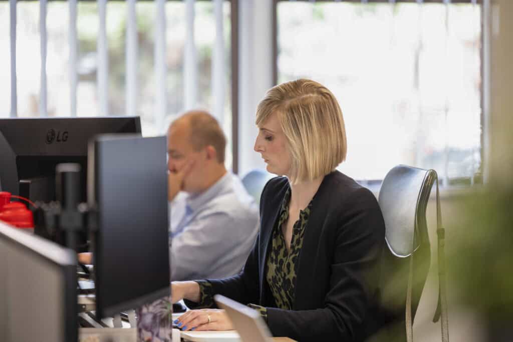Side profile view of Helen Hunt from GLX Advisory who is working at her desk. Steven Hemp from GLX Advisory can be seen behind her blurred in the background.