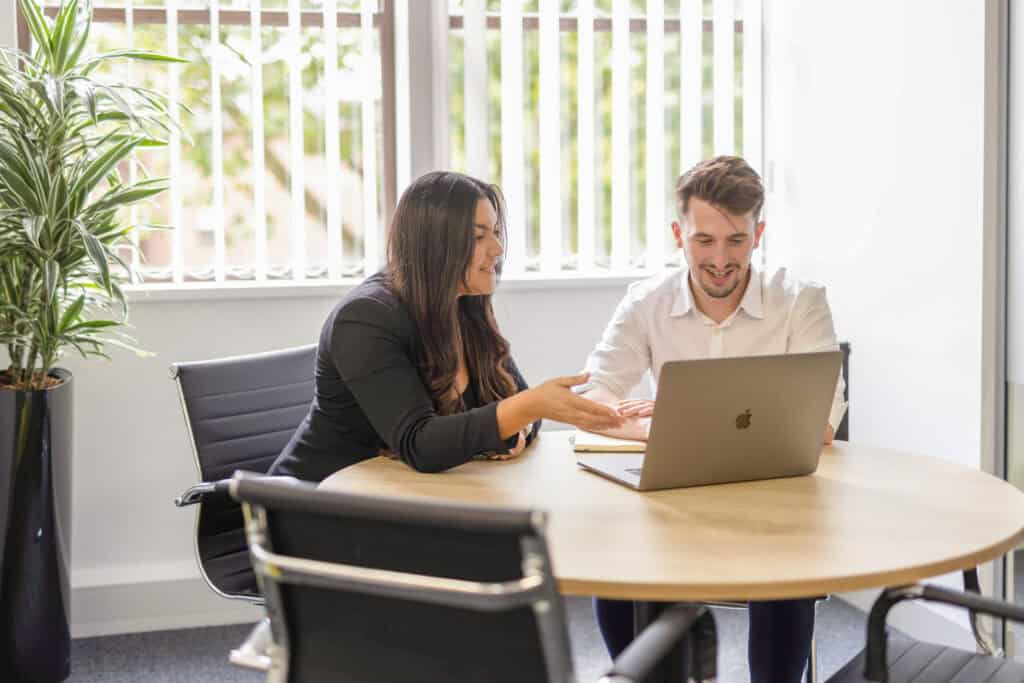 Melissa Porter and Edward Gilbert sit as a table in the meeting room discussing work on a laptop