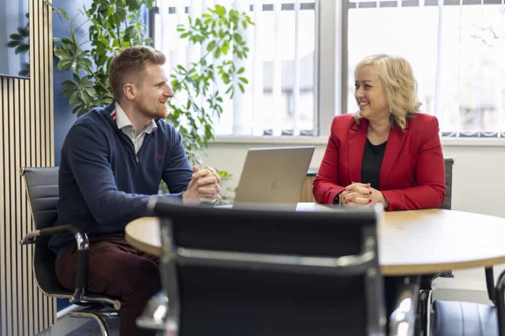 Will Moy, manager of GLX Accounting wears a blue quarter-zip jumper as he sits at a table talking to Karheena Woodroofe from GLX Advisory who sits opposite wearing a red blazer.