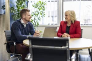 Will Moy, manager of GLX Accounting wears a blue quarter-zip jumper as he sits at a table talking to Karheena Woodroofe from GLX Advisory who sits opposite wearing a red blazer.