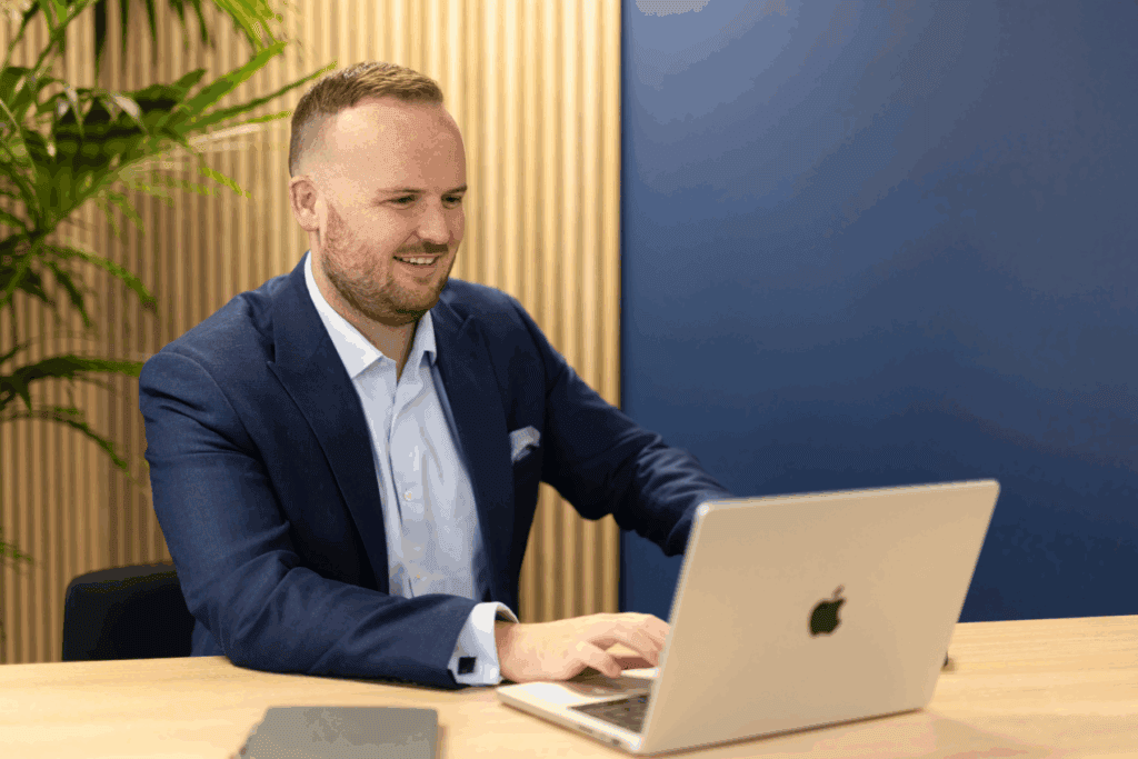 Image for the Making Tax Digital Questions Answered blog is a photo of our Director, Grant Miles, who sits at a desk in a light blue shirt and navy blazer, looking at a laptop.