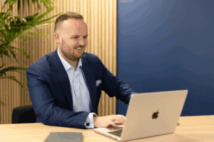 Image for the Making Tax Digital Questions Answered blog is a photo of our Director, Grant Miles, who sits at a desk in a light blue shirt and navy blazer, looking at a laptop.