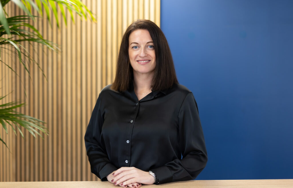 Sara Grodentz - Executive Assistant for GLX stands in front of a wooden slatted wall and a blue wall.