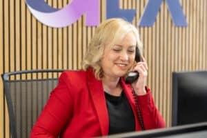 Karheena Woodroffe of GLX Advisory smiles while on the telephone wearing a red blazer and black blouse whilst in front of a slatted wooden wall which features the bottom half go the GLX logo.