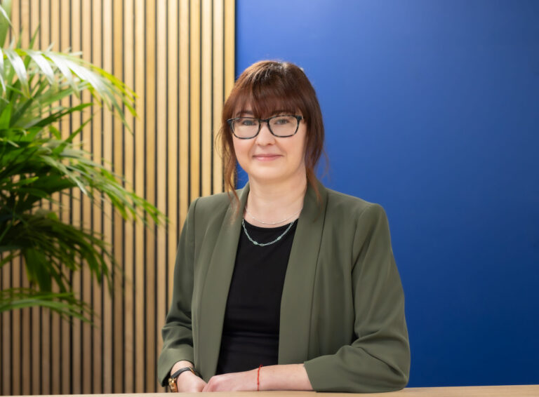 Louise Whistlecraft from the GLX payroll department stands in front of a blue wall which has wood panelling to the left in the background.