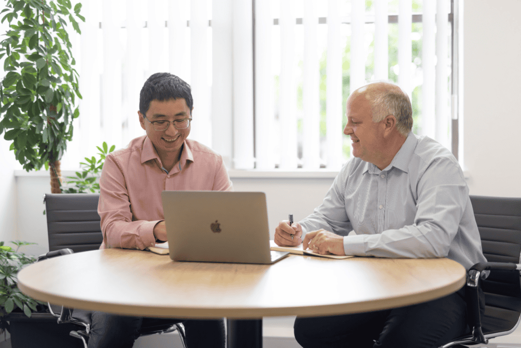 Ke Huang (left) and Duncan Smith (right) from the GLX team sit next to each other at a table. Ke is demonstrating something on a laptop, and Duncan takes notes. There is a window behind them with greenery in the back left corner.
