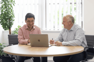 Ke Huang (left) and Duncan Smith (right) from the GLX team sit next to each other at a table. Ke is demonstrating something on a laptop, and Duncan takes notes. There is a window behind them with greenery in the back left corner.