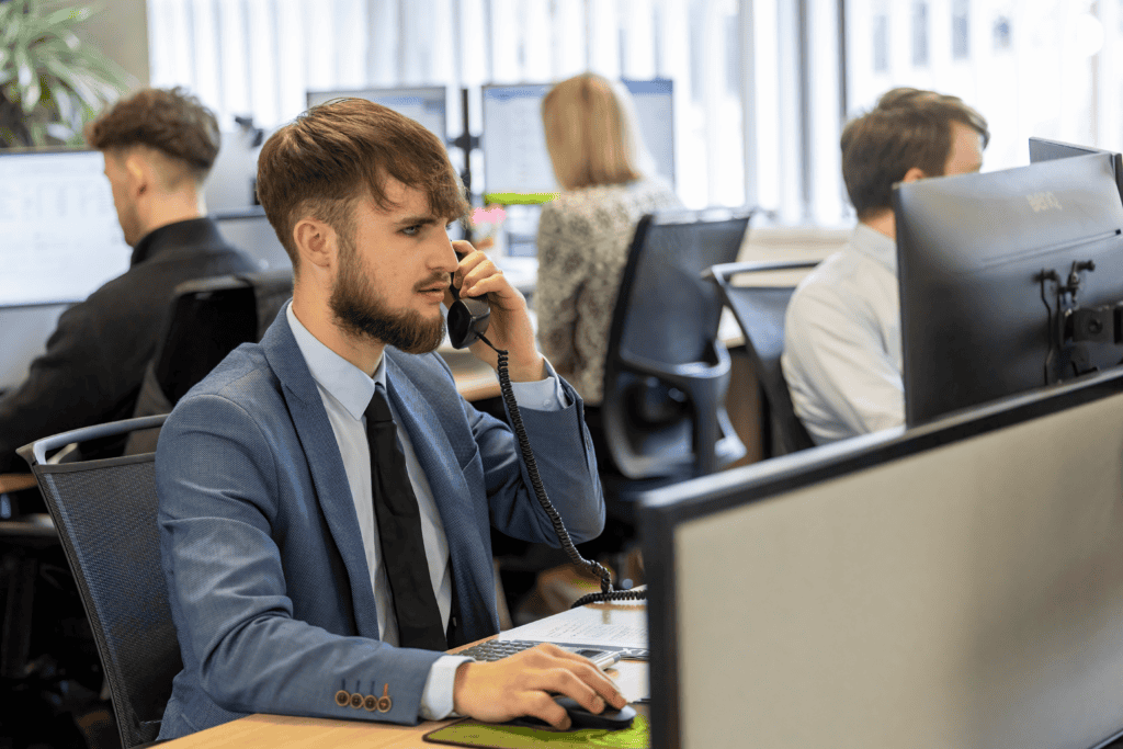 Supporting image for the £3,000 Youth Jobs Grant blog post: Assistant Accountant Adam Mundy from GLX Advisory sits as his desk looking to the right, with his right hand on the mouse, and holding the phone in his left hand while he has a conversation. GLX team members can be seen blurred in the background.
