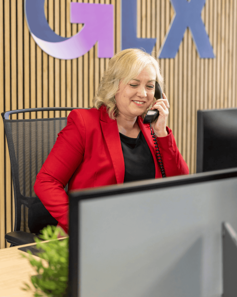 Karheena Woodroffe of GLX Advisory smiles while on the telephone wearing a red blazer and black blouse whilst in front of a slatted wooden wall which features the bottom half of the GLX logo.