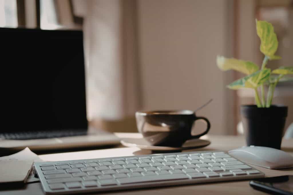 A decorative image of a close up of an apple keyboard with a screen, plant, mouse, and cup blurred in the background. This image support the "What business expenses can I claim working form home" article.