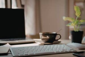 A decorative image of a close up of an apple keyboard with a screen, plant, mouse, and cup blurred in the background. This image support the "What business expenses can I claim working form home" article.