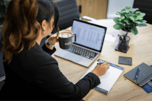 Melissa Porter from GLX sits at a table in front of her laptop with a coffee in her left hand and a pen in her right hand poised over a GLX notepad.