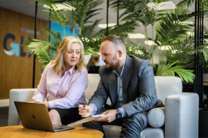 Karheena Woodroffe (left) and Grant Miles (right) from GLX sit on a sofa with a coffee table in front of them which has a laptop on it. Grant holds a notepad and a pen. In the background, the GLX logo wall can be seen blurred.